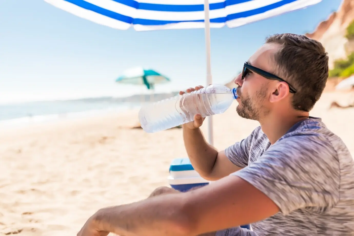 A young man drinks water on the beach under a green solar umbrella.